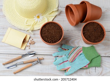 High Angle Shot Of A Group Of Items For Potting And Planting Seeds. Horizontal Format On A Rustic Wooden Potting Table. Items Include, Shovel, Hat, Flower Pots, Soil, Gloves, Seed Packet And More. 