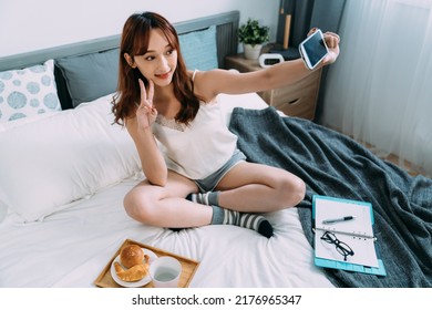 High Angle Shot Happy Cute Asian Girl Holding Mobile Phone Is Showing Victory Sign While Taking Selfie With Breakfast Served On Bed In A Bright Bedroom At Home.