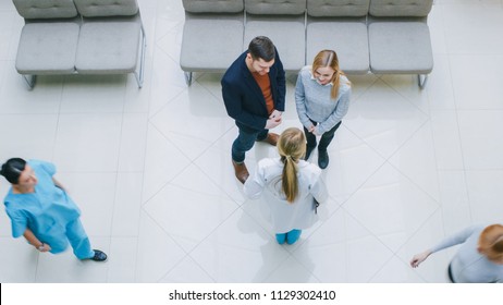 High Angle Shot In The Hospital Lobby, Young Couple Talks Meets With  Female Physician / Doctor / Nurse, They Have Discussion. New, Clean, Modern Hospital With Friendly And Professional Personnel.