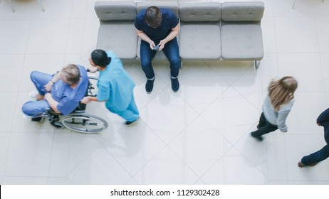High Angle Shot in the Hospital Lobby, Young Man Waits for Results while Sitting and Using Mobile Phone, Doctors, Nurses and Patients Walk Past Him. Clean, New Hospital.