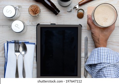 High Angle Shot Of A Man Holding His Glass Of Beer On A Restaurant Table With A Table Computer Menu For Ordering. Only The Man's Hand And Arm Are Visible.