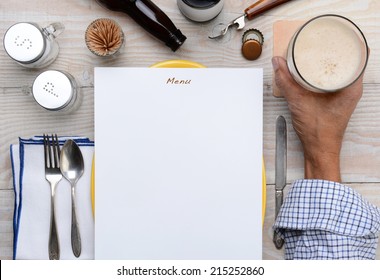 High Angle Shot Of A Man Holding His Glass Of Beer On A Restaurant Table With A Blank Menu In Front Of Him. Only The Man's Hand And Arm Are Visible.