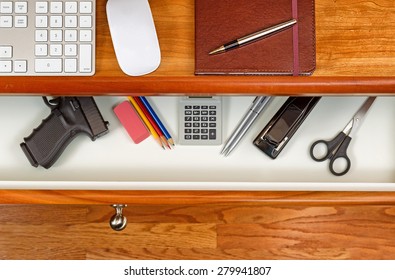 High angle shot of open desk drawer with concealed personal weapon inside. Cherry desktop has computer keyboard, mouse and executive notepad with pen. 