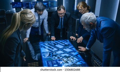 High Angle Shot of Team of Government Intelligence Agents Talking while Standing Around Digital Touch Screen Table and Tracking Suspect. Satellite Surveillance Operation in the Monitoring Room.