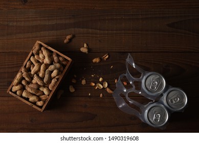 High Angle Shot Of Three Beer Cans In A Plastic 6-pack Holder And A Wood Box Of Peanuts. Used Shells Litter The Rustic Dark Wood Table.