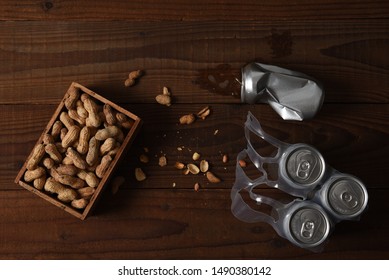 High Angle Shot Of Three Beer Cans In A Plastic 6-pack Holder A Crushed Can And A Wood Box Of Peanuts. Used Shells Litter The Rustic Dark Wood Table.