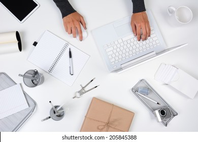 High angle shot of a woman working from her home office. Only her hands are visible as she types on her laptop computer. The white desk is neat with white and silver items.