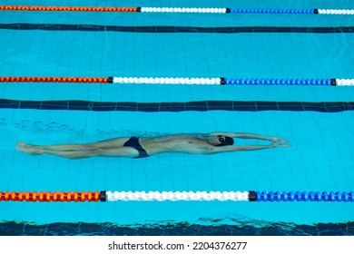 High Angle Side View Of Caucasian Male Swimmer At Swimming Pool, Swimming Under Water On His Back, Wearing Black Swimming Cap And Swimming Goggles