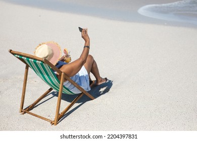 High angle side view of a senior African American woman on a beach in the sun, sitting in a deckchair, wearing a sun hat, holding a cocktail and taking selfies with her smartphone - Powered by Shutterstock