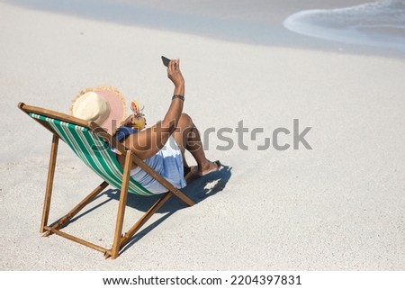 High angle side view of a senior African American woman on a beach in the sun, sitting in a deckchair, wearing a sun hat, holding a cocktail and taking selfies with her smartphone