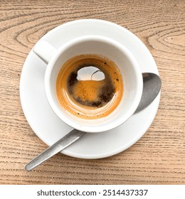 High angle square view over a delicious and beautiful expresso coffee in a white cup with a spoon. Beautiful brown foam color and aroma. Wooden table background.