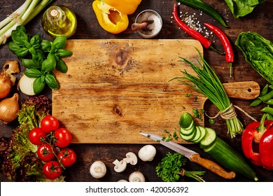 High Angle Still Life View of Knife and Wooden Cutting Board Surrounded by Fresh Herbs and Assortment of Raw Vegetables on Rustic Wood Table