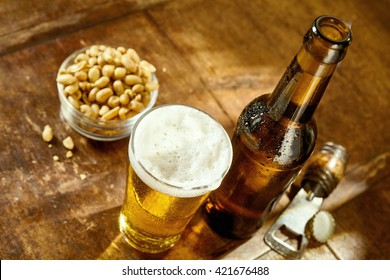 High Angle Still Life View of Glass of Cold Ale on Rustic Wooden Table Beside Bottle of Beer, Bowl of Peanuts and Bottle Cap Opener