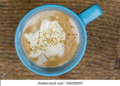 High Angle Still Life View Of Frothy Cappuccino Coffee Served In Blue Mug And Topped With Dollop Of Whipped Cream And Sprinkled With Cinnamon Sugar