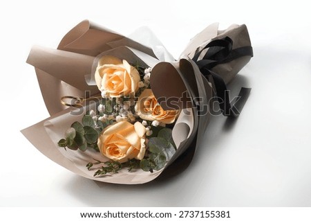 High angle studio shot of an elegant bouquet featuring three champagne roses nestled with white baby's breath and green eucalyptus foliage. The arrangement is professionally wrapped in layered muted brown and white kraft paper, finished with a stylish black ribbon bow. Set against a clean white background, this minimalist and romantic floral composition is ideal for Valentine's Day, Mother's Day, anniversaries, or luxury gift concepts.