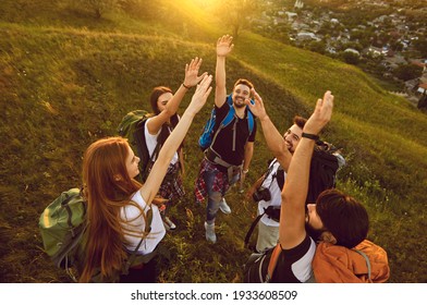 High angle of team of happy young tourists reaching out for high five while hiking on green grassy hill in the evening. From above of group of smiling people with backpacks having fun on trekking tour - Powered by Shutterstock