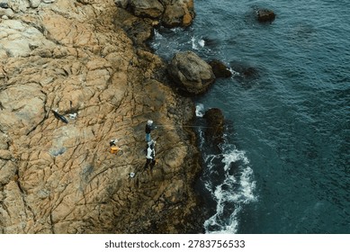High angle, top-down view of local fishermen standing on a rugged, craggy rocky coastline as deep blue ocean waves crash against the shore in Busan, South Korea.