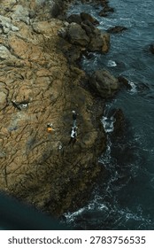 High angle, top-down view of local fishermen standing on a rugged, craggy rocky coastline as deep blue ocean waves crash against the shore in Busan, South Korea.