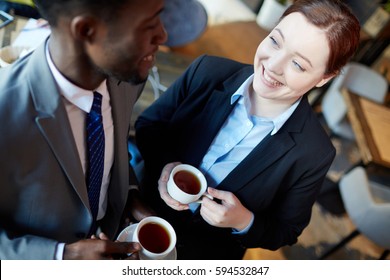 High Angle Of Two Business People Interacting During Coffee Break: Young Woman Talking To African-American Colleague Smiling And Holding Cups With Tea