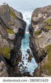 High angle vertical photograph of dramatic sea cliffs along the Wild Atlantic Way in Ireland. Rough Atlantic Ocean waves and turquoise water surge through a narrow gorge between steep rock walls covered with patches of grass and moss. The scene shows rugged geology, scattered boulders in white foaming surf and an overcast sky above the open sea, conveying the wild character of the Irish coastline.