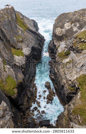 High angle vertical photograph of dramatic sea cliffs along the Wild Atlantic Way in Ireland. Rough Atlantic Ocean waves and turquoise water surge through a narrow gorge between steep rock walls covered with patches of grass and moss. The scene shows rugged geology, scattered boulders in white foaming surf and an overcast sky above the open sea, conveying the wild character of the Irish coastline.