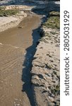 A high angle vertical shot of a shallow freshwater stream carving its way through a sandy beach with green patches of seaweed on the banks, emptying into the sea