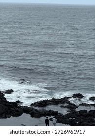 High angle vertical view of vast grey ocean surface and two people standing on black rocks