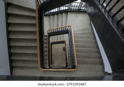 High angle vertigo perspective looking down a geometric square spiral staircase with a wooden handrail and terrazzo steps inside a historic modernist university building.