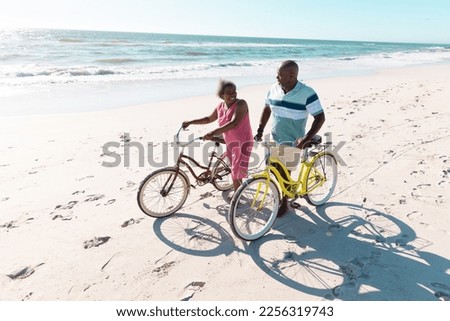 High angle view of african american senior couple with bikes talking and walking at beach in summer. Copy space, travel, unaltered, vacation, love, together, retirement, nature and active lifestyle.