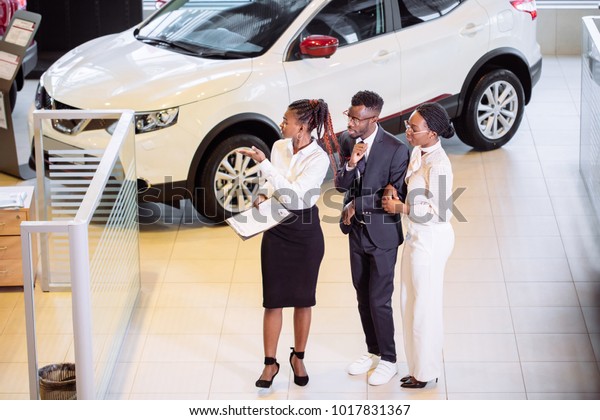 High\
angle view of african car saleswoman standing at dealership telling\
about features of car to african couple\
customers