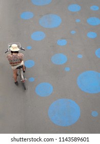 High Angle View Of An Anonymous Woman  With A White Hat, Ridding A Bycicle, In An Asphalt Or Concrete Road, Full Of Blue Rounds Of Street Art, In Paris, France, Europe.