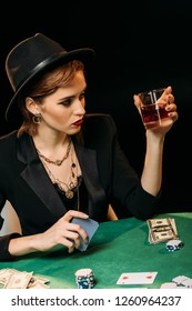 High Angle View Of Attractive Girl In Jacket And Hat Holding Glass Of Whiskey While Playing Poker At Table In Casino