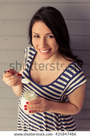 High angle view of beautiful pregnant woman eating yogurt, looking at camera and smiling, standing against gray wall