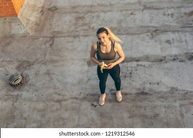 High Angle View Of A Beautiful Young Woman Standing On A Building Rooftop Terrace, Taking A Workout Break And Eating Banana