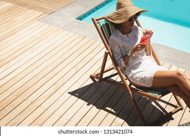 High Angle View Of Beautiful Young African American Woman With Cocktail Glass Relaxing On Sun Lounger In Backyard On A Sunny Day. She Seems To Be On Vacation