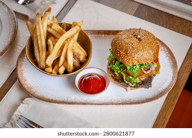 High angle view of beef burger and french fries plate. High angle shot of a gourmet beef cheeseburger, thick cut french fries, and ketchup bowl on a ceramic platter.
