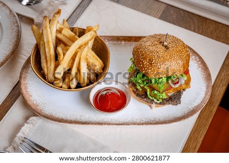 High angle view of beef burger and french fries plate. High angle shot of a gourmet beef cheeseburger, thick cut french fries, and ketchup bowl on a ceramic platter.