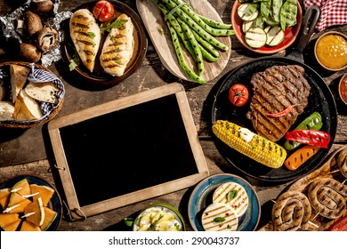 High Angle View Of Blank Chalkboard Amongst Grilled Meal Of Steak, Chicken And Vegetables Spread Out On Rustic Wooden Table At Barbeque Party