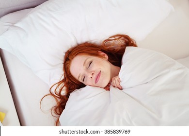 High Angle View Of Blissful Looking Young Woman With Long Red Hair Smiling And Lying Snuggled Up In Comfortable Bed With White Linens In Luxury Hotel Bedroom