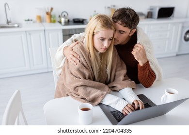 high angle view of blonde woman using laptop while boyfriend hugging her near cups on table