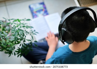 High Angle View Of Boy Wearing Headphone And Reading Books At Home. He Is Sitting Cross Legged On Floor And Feel Comfortable. Child Self Learning And Studying During Covid-19 Background.