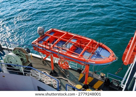 High angle view of a bright orange rescue boat with an outboard motor mounted on a ship deck above turquoise sea water, featuring maritime safety equipment and lifebuoy.