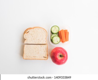 High Angle View Of Brown Bread Sandwich, Red Apple, Cucumber And Carrots On White Background - Healthy Packed Lunch Concept