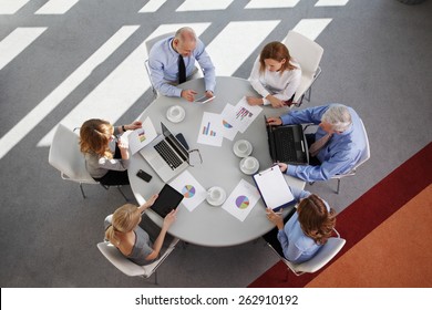 High Angle View Of Business People Discussing In A Meeting While Sitting At Conference Table. Using Digital Tablet And Computer While Analyzing Financial Data.  Shot Taken From Directly Above.