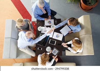 High angle view of business team sitting around the desk and shaking hands. Businesswomen and businessmen working with laptop and digital tablet while making business deal. 