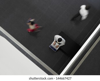 High Angle View Of Businessman Using Laptop With People Walking By In Corridor