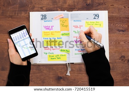 High Angle View Of A Businessperson With Mobile Phone Writing Schedule In Diary
