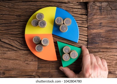 High Angle View Of Businessperson's Hand Taking Piece With Money From Pie Chart