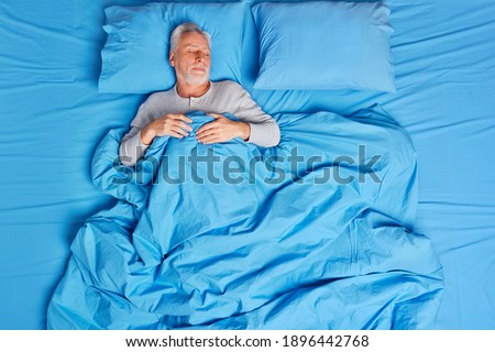 High angle view of calm bearded grey haired senior man sleeps peacefully in bed enjoys pleasant dreams feels tired after hard day lives alone poses on soft blue pillow. Early morning concept