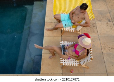 High Angle View Of A Caucasian Couple Wearing Beachwear Sitting On Chairs Sunbathing In A Garden Beside A Swimming Pool, The Woman Wearing A Sun Hat And Using A Laptop, The Man Using A Smartphone